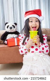 Smiling Girl In Santa Helper Hat And Holding Glass Drinking Water With Teddy Bear Sitting On Isolated White Background.