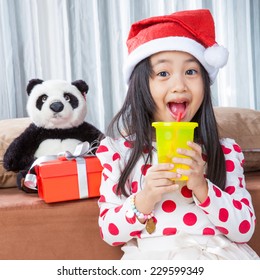 Smiling Girl In Santa Helper Hat And Holding Glass Drinking Water With Teddy Bear Sitting On Isolated White Background.