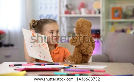Smiling girl showing drawings teddy bear, playroom leisure, imaginary friend