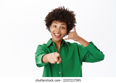 Smiling girl showing phone and pointing at you, call us gesture, callcenter advertisement, contact us, standing over white background