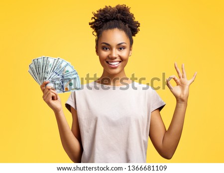 Smiling girl shows money cash and excellent sign, demonstrates that everything is fine. Photo of african american girl wears casual outfit on yellow background. Emotions and pleasant feelings concept.