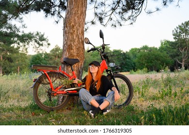 Lächelndes Mädchen sitzend in der Nähe von rotem Elektro-Fahrrad auf grünem Gras mit Strauß von wilden Blumen.Erholung genießen nach Radfahren bei Sonnenuntergang Sommertag. Erholung, Menschen, gesundes Lebensstil
