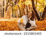 Smiling girl sits in the autumn foliage on the ground of the forest with the yellow leaves