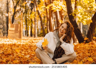 Smiling girl sits in the autumn foliage on the ground of the forest with the yellow leaves