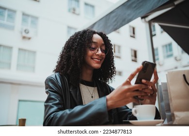 Smiling girl sitting at a cafe terrace while she is sending a text message on her phone to friends - Shutterstock ID 2234877813