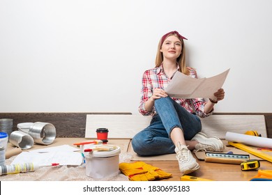 Smiling Girl Sitting On Floor And Holding Paper Blueprint. Home Remodeling And House Interior Redesign. Construction Tools And Materials For Building. Young Attractive Woman Wearing Casual Clothes