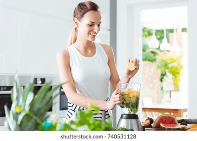 Smiling girl squeezing lemon juice into blender with water and sliced fruit while preparing smoothie - Powered by Shutterstock