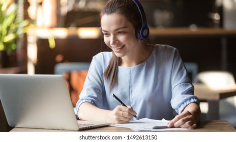 Estudiante sonriente usa audífono inalámbrico en línea con profesora de skype, joven feliz aprende el idioma escucha conferencias ver webinar notas escribir mirar el sitio de una laptop en un café, educación distante