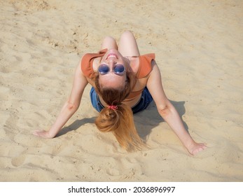 Smiling girl in sunglasses sits on the sand with her head thrown back