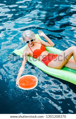 smiling girl in swimsuit relaxing on green inflatable mattress in swimming pool with watermelon