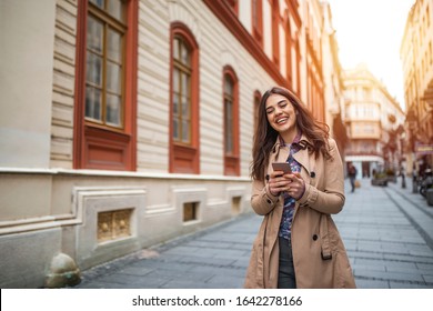 Smiling Girl Texting Outside. Caucasian Woman Female Using Iphone Cellphone Smartphone Walking Streets. Portrait Of Young Happy Woman Walking In City And Using Smartphone. Girl Browse The Internet