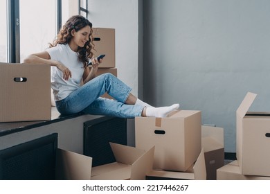 Smiling Girl Using Smartphone Apps, Sitting On Window Sill With Boxes On Relocation Day, Happy Female Home Buyer Choosing Moving Service, Or Looking For Interior Design Ideas, Planing Home Renovation.