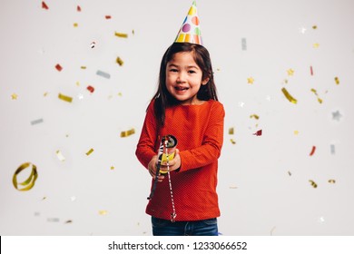 Smiling Girl Wearing Cone Cap Bursting A Party Popper. Little Girl Holding A Party Popper With Confetti Flying All Around.