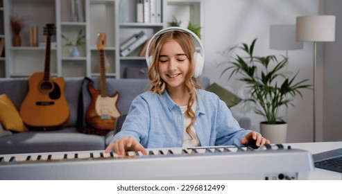 Smiling girl wearing white headphone learning and playing piano music in the living room at home studio. Little female studying online. - Powered by Shutterstock