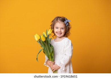 Smiling Girl In White Dress On Yellow Background. Cheerful Happy Child With Tulips Flower Bouquet