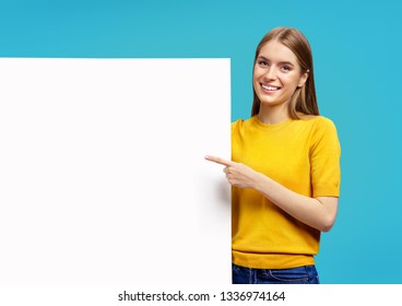 Smiling girl with white empty board, pointing away, on blue background. Copy space for your text.