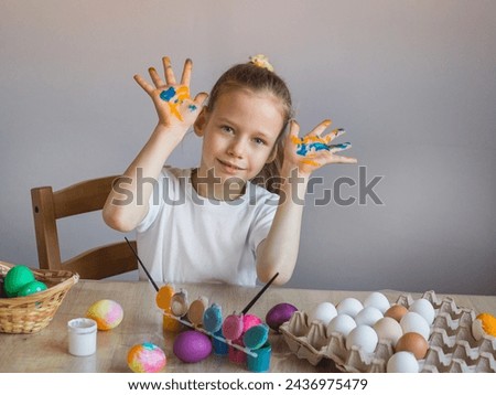 
A smiling girl in a white T-shirt shows her palms covered in paint stained with eggs. Easter. Spring. Painted eggs. Multi-colored children's lodons. Children's hands in paint. 