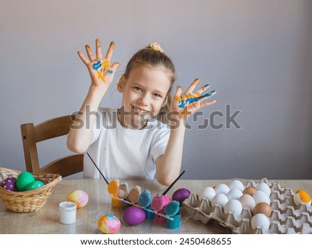
A smiling girl in a white T-shirt shows her palms covered in paint stained with eggs. Easter. Spring. Painted eggs. Multi-colored children's lodons. Children's hands in paint. Happy child celebrating