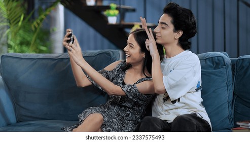 Smiling girlfriend sitting on sofa taking funny faces selfie with teen boy using mobile phone at indoor home. Happy Indian teenager girl making self photos record vlog on smartphone enjoy fun together