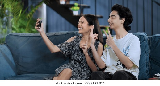 Smiling girlfriend sitting on sofa taking funny faces selfie with teen boy using mobile phone at indoor home. Happy Indian teenager girl making self photos record vlog on smartphone enjoy fun together - Powered by Shutterstock