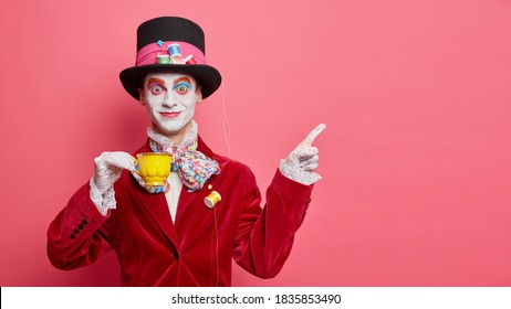 Smiling glad hatter in fashionable costume has aristocratic manners drinks tea and indicates at copy space isolated over rosy background. Man has image of classic tale character for halloween