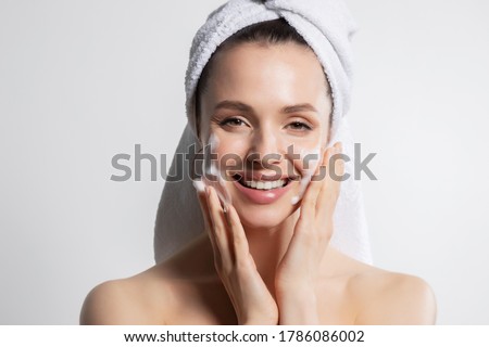 Smiling glad woman applying cosmetic foam portrait. Cheerful laughing millennial washing on face looking at camera. Lovely brunette in head bath towel with attractive appearance. Skincare spa