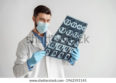 Smiling glad young caucasian man doctor in face mask and medical gown analyzing lungs x-ray MRI scan viewing good treatment results. Portrait isolated on white background. Healthcare and medicine
