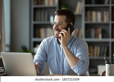 Smiling good-looking businessman in glasses sit at workplace desk in modern office room using wireless device holding smartphone talking to client, solve issues remotely. Business conversation concept