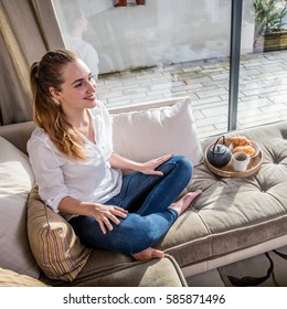 smiling gorgeous young woman with poney tail sitting on a comfortable sofa with her breakfast, tea and croissants in a comfortable home with view on the terrasse