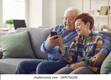 Smiling Grandfather And Grandson Sitting On Sofa And Having Fun. Little Child Showing Grandpa How To Use Social Media App On Smartphone. Happy Elderly Man Learning To Make Video Call On Mobile Phone