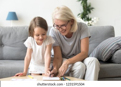 Smiling Grandmother And Little Kid Granddaughter Drawing On Paper With Colored Pencils Together, Caring Granny Teaching Grandchild Having Fun Playing At Home, Grandma And Creative Child Activity