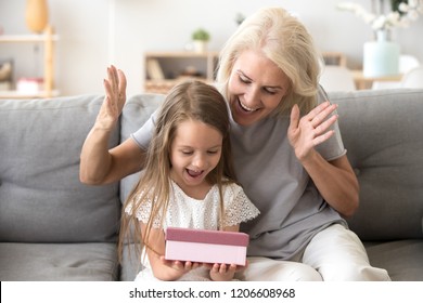 Smiling Grandmother Making Surprise To Cute Little Granddaughter Giving Gift Box, Excited Small Girl Get Birthday Present From Granny, Happy Older And Younger Generation Celebrate At Home Together