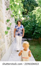 Smiling Grandmother Walking Behind Her Little Granddaughter Near The Stone Wall In The Garden