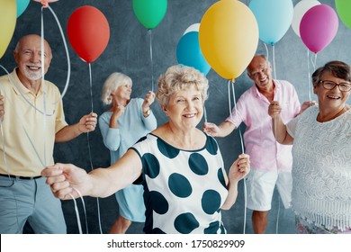 Smiling Grandmother Wearing A White Blouse With Black Dots And Holding Colorful Balloons During New Year's Eve Party With Senior Friends