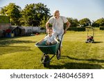 Smiling grandson sitting in wheelbarrow and his granddad is pushing it. Family having fun while gardening in backyard.