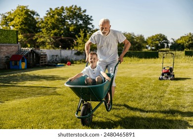 Smiling grandson sitting in wheelbarrow and his granddad is pushing it. Family having fun while gardening in backyard. - Powered by Shutterstock