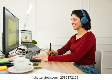 Smiling graphic designer using a pen tablet and computer while wearing headphones, working remotely in a well-lit home office