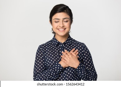 Smiling grateful Indian young woman holding hands on chest isolated on grey studio background, pleased beautiful girl with closed eyes feeling love, gratitude, appreciation, thanking fate