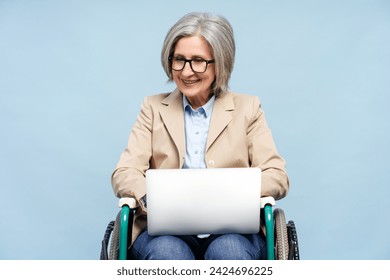 Smiling gray haired senior woman using laptop computer, typing, working online sitting in wheelchair isolated on blue background. Freelance job concept - Powered by Shutterstock