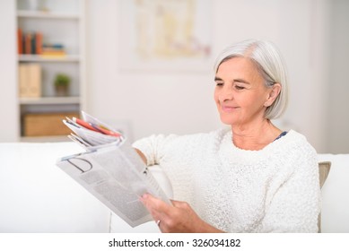 Smiling Gray-Haired Senior Lady Reading A Magazine At The White Couch In The Living Room.