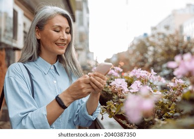 Smiling grey haired mature Asian woman in stylish shirt uses cellphone standing on outdoors terrace decorated with pink flowers