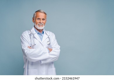 Smiling grey-haired male doctor, physical, therapist wearing white medical gown with stethoscope on shoulders stands with arms crossed isolated on blue, copy space. Healthcare and medicine - Powered by Shutterstock