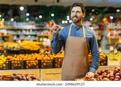 Smiling grocery store worker standing in produce section pointing at something. Employee wearing apron, indicating something to customers or colleagues, surrounded by fresh fruits and vegetables - Powered by Shutterstock