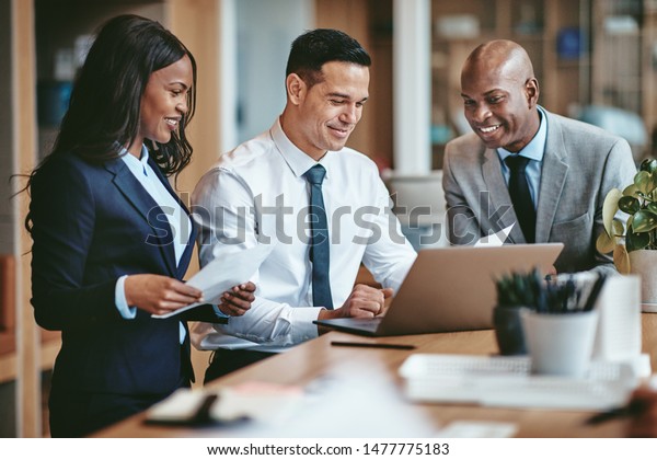 Smiling group\
of diverse businesspeople going over paperwork together and working\
on a laptop at a table in an\
office