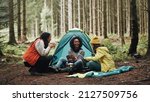 Smiling group of diverse young female friends sitting at their forest campsite drinking coffee and reading a trail map