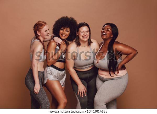 Smiling group of women in different size\
standing together in sportswear against brown background. Diverse\
group women looking at camera and\
laughing.
