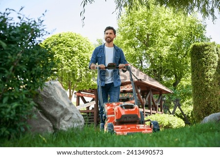 Smiling guy with beard trimming overgrown green lawn with electric mower in garden. Low angle view of happy male gardener in shirt using lawn trimmer, while landscaping. Concept of seasonal work.