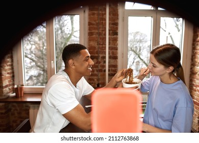 Smiling Guy Feeding Girlfriend Noodles During Making Eat Fast Food Video Challenge On Smartphone. Video Blogging. Young Multiethnic Couple Of Generation Z Creating Content For Social Networks At Home