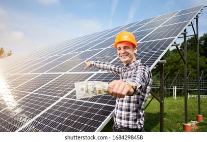 Smiling Guy With Hundred Dollar Bill In Hand Shows At Station Of Solar Panels In Which The Sun's Rays Are Reflected. Business And Generation Concept. Green Ecological Power Energy Generation.