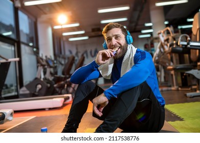A smiling guy sits after a workout in the gym, listens to music on headphones and eats a protein bar